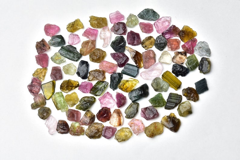 May include: A collection of rough, natural tourmaline gemstones in various colors, including green, pink, black, and yellow. The stones are arranged in a circular pattern on a white background.