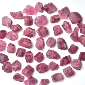 May include: A collection of raw, pink tourmaline gemstones. The rough, irregular stones vary in size and shape, displaying a range of pink hues. The gemstones are scattered on a white surface, creating a natural and earthy aesthetic.