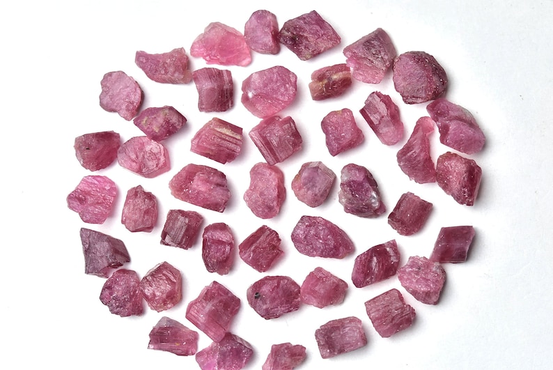 May include: A collection of raw, pink tourmaline gemstones. The rough, irregular stones vary in size and shape, displaying a range of pink hues. The gemstones are scattered on a white surface, creating a natural and earthy aesthetic.