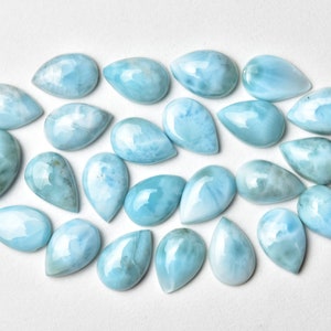 May include: A collection of 20 teardrop-shaped, light blue, iridescent gemstone cabochons. The cabochons are arranged in a semi-circle on a white background.