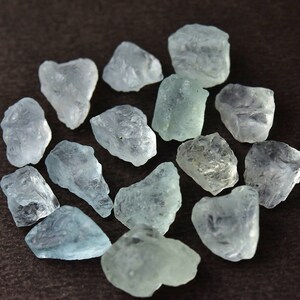 Puede incluir: Un conjunto de piedras preciosas de aguamarina en bruto. Las piedras, de varios tama&ntilde;os y formas, exhiben un tono azul verdoso p&aacute;lido. Est&aacute;n dispuestas en un patr&oacute;n circular sobre un fondo marr&oacute;n oscuro, mostrando su textura cristalina natural.