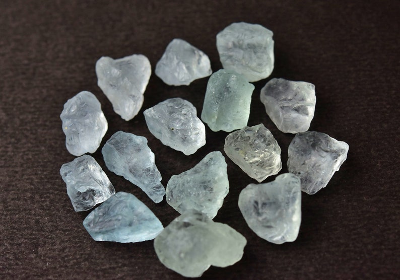 Puede incluir: Un conjunto de piedras preciosas de aguamarina en bruto. Las piedras, de varios tama&ntilde;os y formas, exhiben un tono azul verdoso p&aacute;lido. Est&aacute;n dispuestas en un patr&oacute;n circular sobre un fondo marr&oacute;n oscuro, mostrando su textura cristalina natural.