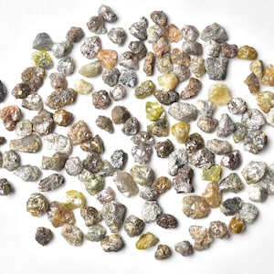 May include: A collection of small, rough, grey, brown, and yellow diamonds. The diamonds are scattered on a white surface.