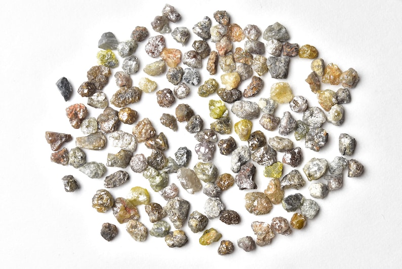 May include: A collection of small, rough, grey, brown, and yellow diamonds. The diamonds are scattered on a white surface.