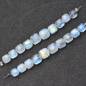 May include: Two strands of moonstone beads. Each bead is a square shape with rounded edges. The beads have a translucent, milky white color with flashes of blue and iridescent hues. The beads are strung on thin silver wires, ready for jewelry making.