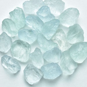 Puede incluir: Un conjunto de gemas de aguamarina en bruto, de color azul claro. Los cristales naturales, de tama&ntilde;os y formas variadas, presentan un aspecto transl&uacute;cido. Las piedras est&aacute;n dispuestas sobre una superficie blanca, resaltando su color azul p&aacute;lido y textura natural.