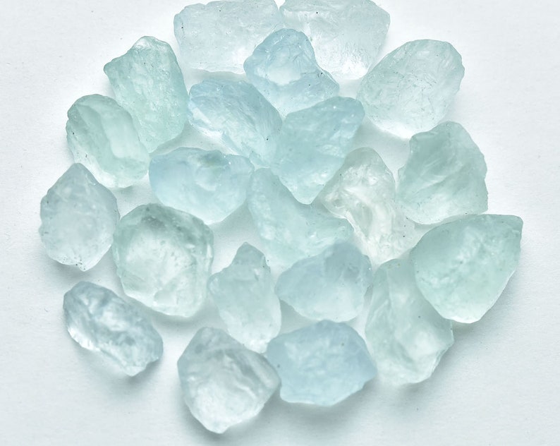 Puede incluir: Un conjunto de gemas de aguamarina en bruto, de color azul claro. Los cristales naturales, de tama&ntilde;os y formas variadas, presentan un aspecto transl&uacute;cido. Las piedras est&aacute;n dispuestas sobre una superficie blanca, resaltando su color azul p&aacute;lido y textura natural.