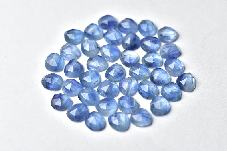 Puede incluir: Un conjunto de gemas facetadas de color azul claro. Cada piedra tiene una forma triangular redondeada con m&uacute;ltiples facetas que capturan la luz. Las gemas est&aacute;n dispersas sobre una superficie blanca, creando un efecto brillante. Ideal para la creaci&oacute;n de joyas.