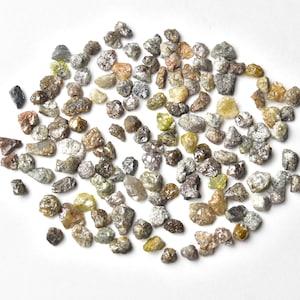 May include: A collection of small, rough, grey, brown, and green diamonds. The diamonds are scattered on a white surface.