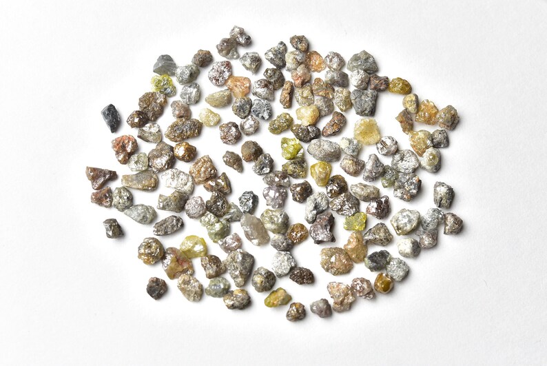 May include: A collection of small, rough, grey, brown, and green diamonds. The diamonds are scattered on a white surface.