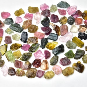 May include: A collection of rough, natural tourmaline crystals in various colors, including green, pink, black, and yellow. The crystals are scattered on a white background.
