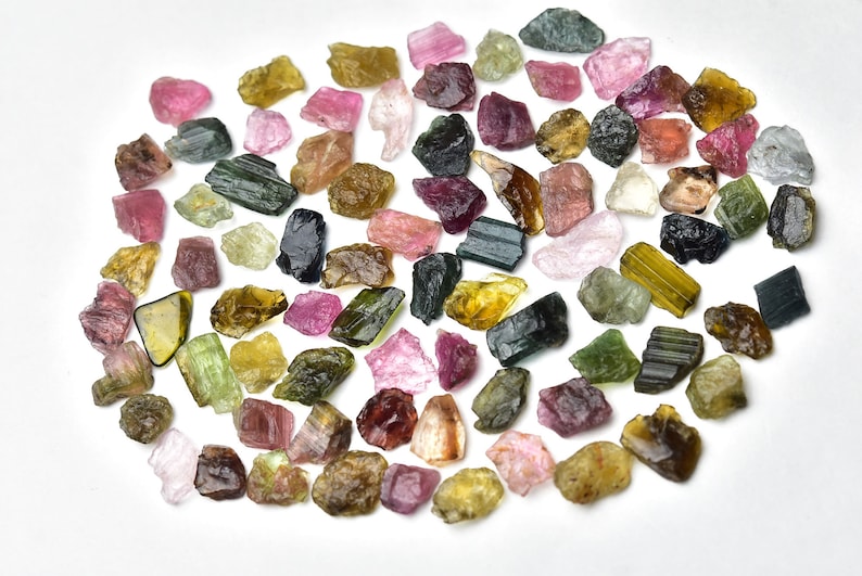 May include: A collection of rough, natural tourmaline crystals in various colors, including green, pink, black, and yellow. The crystals are scattered on a white background.