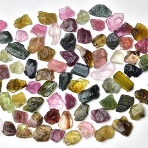 May include: A collection of rough, natural tourmaline gemstones in various colors, including green, pink, brown, and black. The gemstones are scattered on a white background.