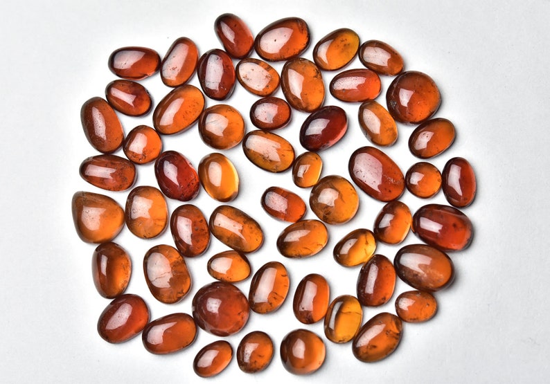 May include: A collection of 40+ oval-shaped, translucent, orange-brown gemstones. The stones are arranged in a circle on a white background.
