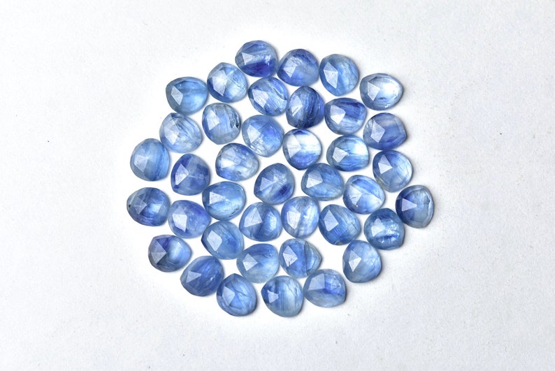 Puede incluir: Colecci&oacute;n de gemas facetadas de color azul claro. Cada piedra tiene una forma redondeada con m&uacute;ltiples facetas, creando un efecto brillante. Las gemas est&aacute;n dispersas sobre una superficie blanca, mostrando su color y claridad.