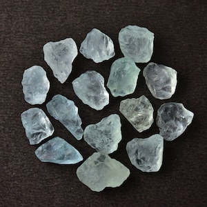Puede incluir: Una colecci&oacute;n de piedras preciosas de aguamarina azul claro en bruto dispuestas en un patr&oacute;n circular. Las piedras tienen una calidad transl&uacute;cida y formas variadas, sobre un fondo marr&oacute;n oscuro.