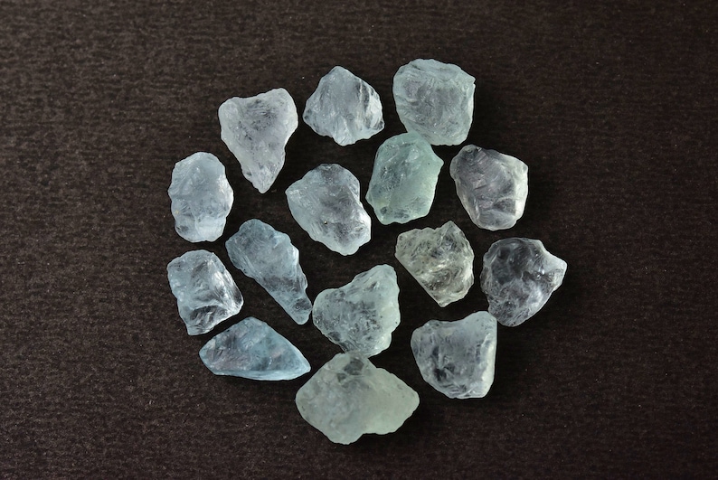 Puede incluir: Una colecci&oacute;n de piedras preciosas de aguamarina azul claro en bruto dispuestas en un patr&oacute;n circular. Las piedras tienen una calidad transl&uacute;cida y formas variadas, sobre un fondo marr&oacute;n oscuro.