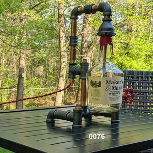 May include: A black metal lamp with a clear glass bottle and a red valve. The bottle has a label that reads "Maker's Mark Whiskey". The lamp is sitting on a black table with a wooden grain pattern.