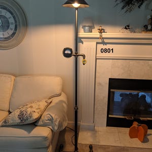 May include: A black floor lamp with a black metal shade and a gold-colored gauge and valve. The lamp is made of industrial pipe and has a black base.