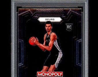 May include: A graded 2023 Prizm Monopoly Victor Wembanyama rookie card. The card features a basketball player in a black and white uniform holding a basketball. The card is encased in a protective holder with the text "2023 PRIZM MONOPOLY VICTOR WEMBANYAMA ROOKIE CARD" at the top.