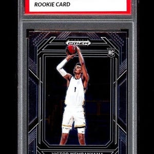 Könnte beinhalten: Eine Rookie-Karte von Victor Wembanyama in weißer Basketballuniform, die einen Ball wirft. Die Karte ist mit "2023 Prizm Draft Picks" und "Rookie Card" beschriftet. Die Karte ist mit MT 10 bewertet.