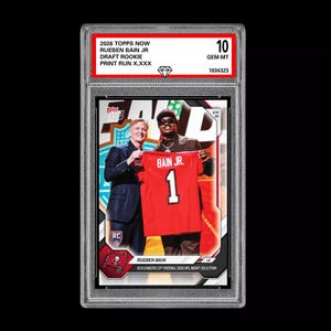 May include: A graded 2026 Topps Now Rueben Bain Jr. Draft Rookie card. The card features a photo of Rueben Bain Jr. holding a red jersey with the number 1. The card is graded a 10 GEM-MT.