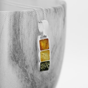 Puede incluir: Un collar con colgante de plata con tres piedras rectangulares de ámbar en tonos amarillos y verdes. El colgante cuelga de una fina cadena de plata. El collar se muestra sobre un fondo borroso y texturizado.