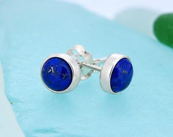 May include: A pair of round stud earrings featuring deep blue lapis lazuli stones set in polished silver bezels. The circular stones display natural dark blue tones with subtle flecks of gold, securely mounted on silver posts with matching backs.
