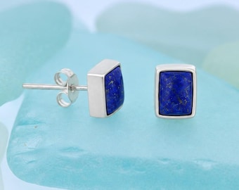 May include: A pair of stud earrings featuring rectangular lapis lazuli stones set in polished silver frames. Each earring has a post back closure. The deep blue stones display natural gold-colored flecks, contrasting with the smooth, bright silver metal settings.