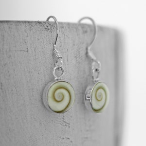 May include: A pair of silver earrings featuring a white and green shell design. The earrings are hanging from a concrete surface.