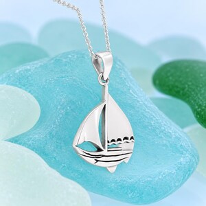 Op de afbeelding: Een zilverkleurige metalen hanger in de vorm van een zeilboot, hangend aan een fijne bijpassende ketting. De hanger heeft een uitgesneden ontwerp met gegraveerde lijnen die golven en zeilen voorstellen, met een gepolijste afwerking en een oogje.