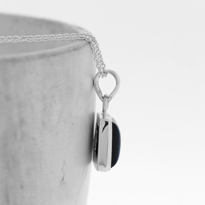 Puede incluir: Un collar de plata con un colgante cuadrado con una piedra preciosa negra.