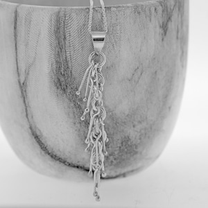 Puede incluir: Un collar de plata con un colgante de cadena larga y delicada y un diseño de borla. El colgante presenta múltiples cuentas pequeñas y redondas y eslabones finos y alargados.