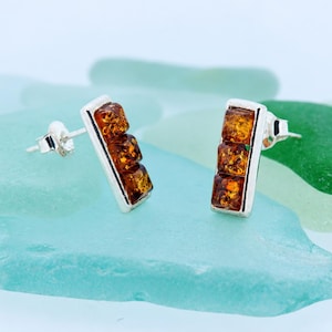 May include: A pair of stud earrings featuring three rectangular, cognac-coloured amber stones set vertically in a polished silver frame. Each earring has a standard post back closure, showcasing the warm, translucent orange-brown tones of the amber.