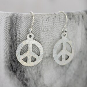 Puede incluir: Un par de pendientes de plata con forma de signo de paz. Los pendientes son simples y elegantes, y serían un gran regalo para cualquiera que ame la paz y el amor.