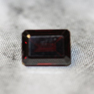 May include: A rectangular, dark red gemstone with a faceted cut. The gem has a deep, rich colour and is set against a textured, off-white background. The gemstone's edges are sharply defined, and the facets catch the light, creating a sparkling effect.