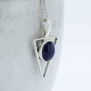 Op de afbeelding: Een zilveren ketting met een hanger met een donkerblauwe edelsteen in een driehoekig frame.