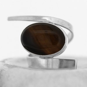 Puede incluir: Un anillo de plata con una piedra ovalada marrón. El anillo tiene un diseño único y moderno con una banda curva.