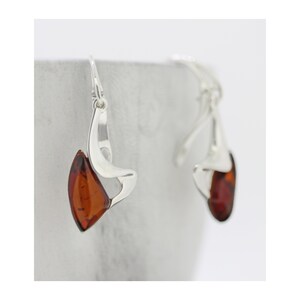 May include: A pair of silver earrings with a teardrop-shaped amber stone. The earrings have a curved, abstract design. The amber stones are a rich, warm brown color, and the silver has a polished finish. The earrings are displayed against a neutral background.