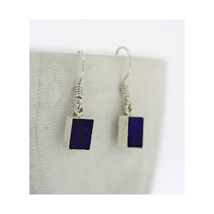 May include: Silver earrings featuring a square, deep blue stone. Each earring has a simple hook design with a coiled detail. The background is a light grey. The earrings are a simple, elegant design.