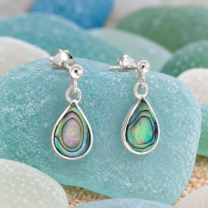 Peut inclure: Une paire de boucles d'oreilles à clous en argent avec des incrustations de coquille d'ormeau en forme de goutte. Chaque boucle présente un sertissage en argent poli encadrant la coquille irisée aux reflets verts, bleus et violets.