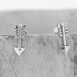 Könnte beinhalten: Ein Paar silberne Pfeil-Ohrstecker. Die Pfeile haben ein detailliertes Design mit einer spitzen Spitze und einem gefiederten Schwanz.