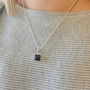 Puede incluir: Un collar de cadena de plata con un colgante cuadrado de piedra negra.