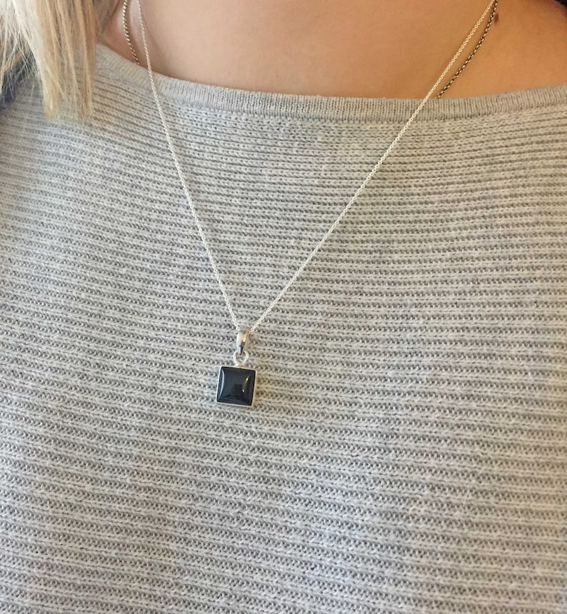 Puede incluir: Un collar de cadena de plata con un colgante cuadrado de piedra negra.