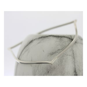 May include: A silver metal headband with a curved, angular design. The headband is displayed on a grey fabric surface.
