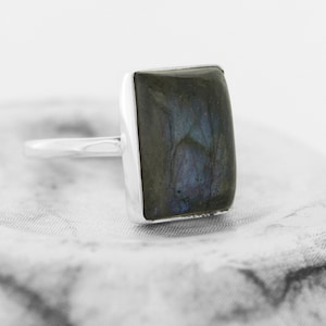 May include: A silver ring featuring a square-shaped labradorite gemstone. The gemstone displays a dark blue and green iridescent sheen.