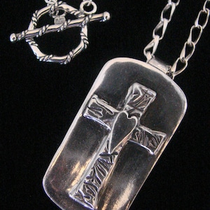 May include: A silver necklace with a toggle clasp and a pendant featuring a cross with a heart in the center. The cross and heart are textured and have a detailed design.