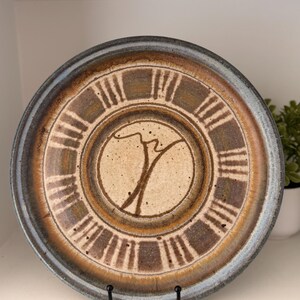 May include: A round ceramic plate with a unique design. The plate features a central circle with a brown tree-like shape and is surrounded by a series of brown rectangles. The outer rim is a darker blue-grey color. The plate is displayed on a black metal stand.