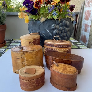 Puede incluir: Una colección de cajas de madera hechas a mano con tapas, de varias formas y tamaños. Las cajas están hechas de madera natural, mostrando la veta y la textura. Algunas cajas tienen elementos decorativos. Un jarrón decorativo gris oscuro con flores coloridas está en el fondo.