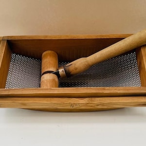 May include: Wooden food mill with a perforated metal screen and a wooden pestle. The mill is rectangular with a wooden frame and a curved wooden base. The pestle is long and cylindrical, with a handle. The mill and pestle are made of light brown wood.